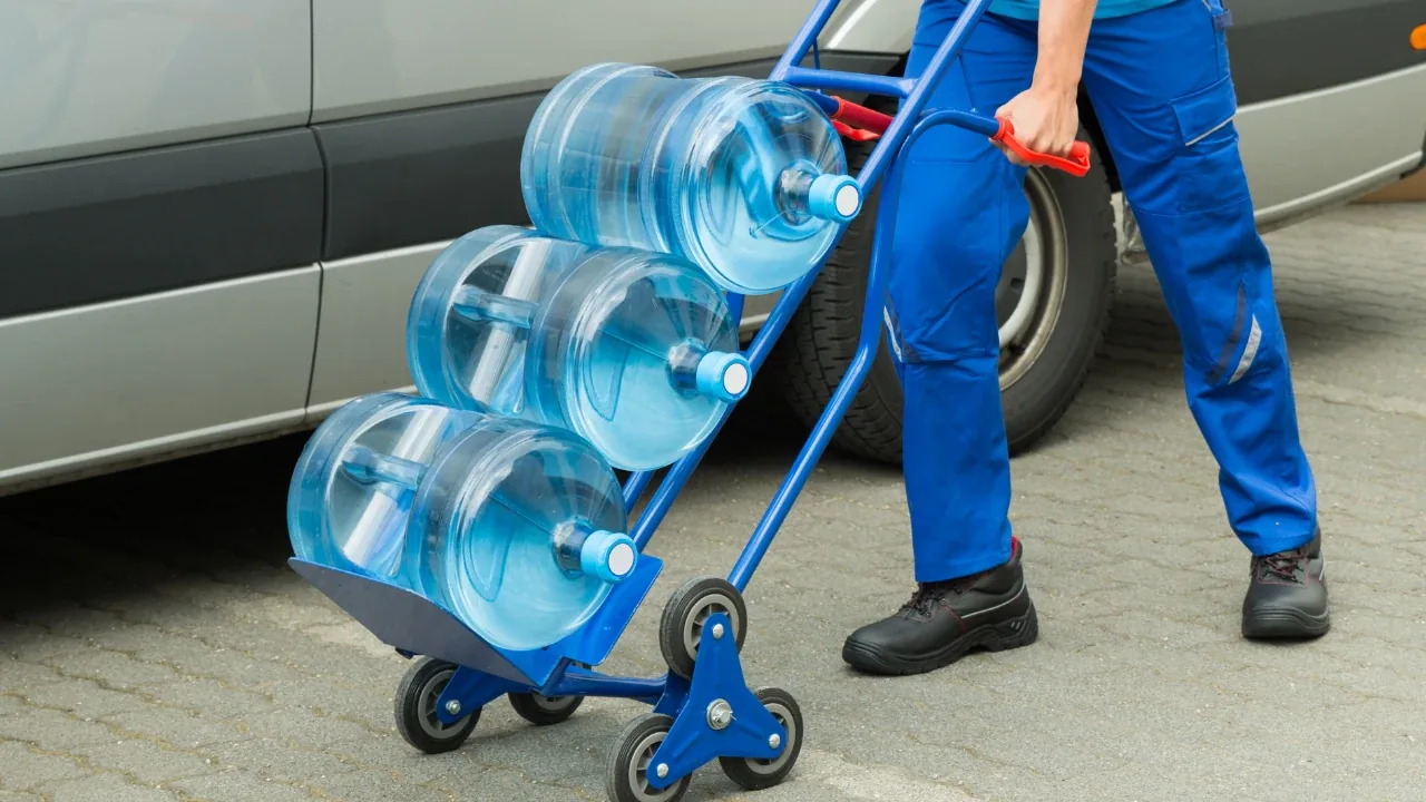 Repartidor trasladando bidones de agua de 20 litros en un carro junto a una camioneta de reparto